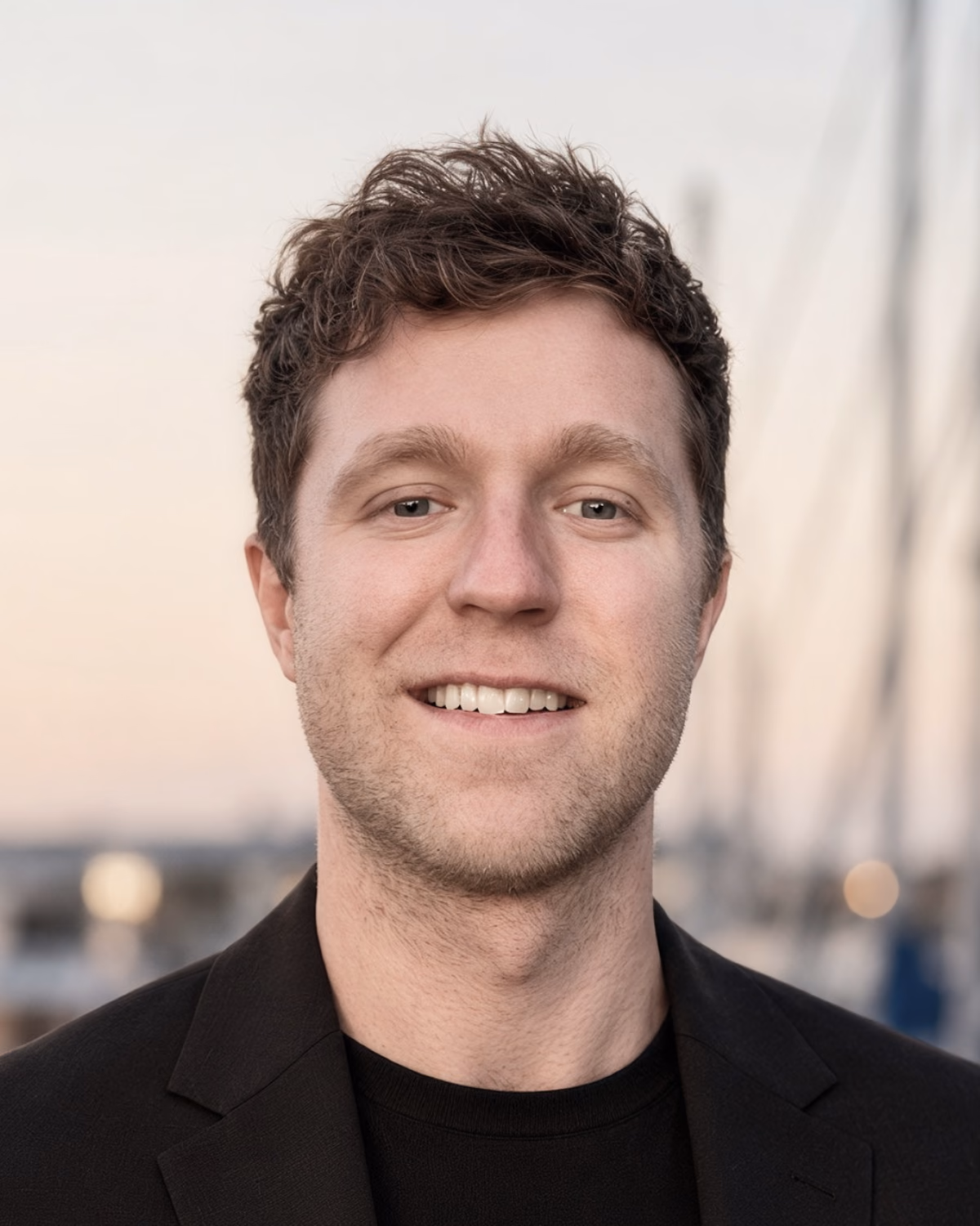 Josh Simpson smiling in a black blazer by a marina.