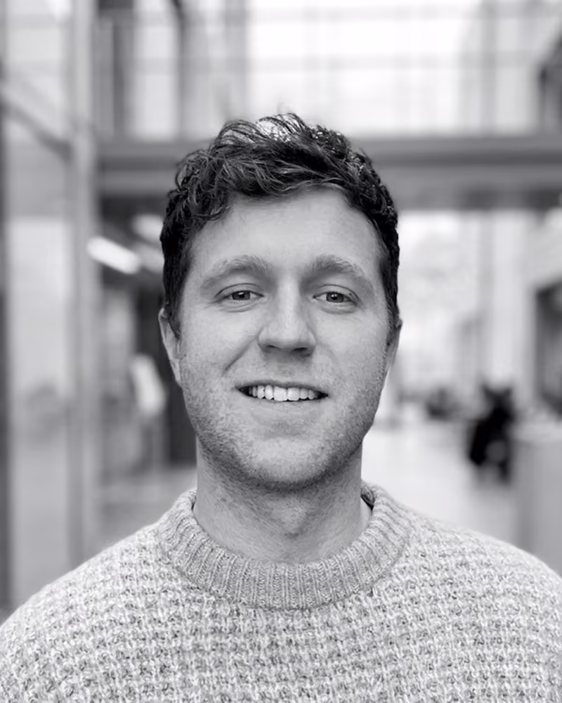 Smiling man with curly hair wearing a knit sweater, standing indoors with blurred background.