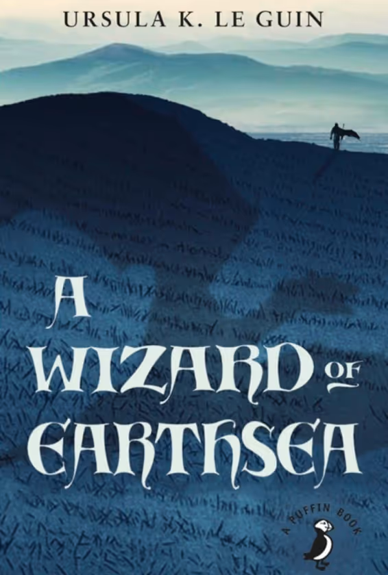 Book cover for 'A Wizard of Earthsea' by Ursula K. Le Guin featuring a person holding a staff walking on dark blue hills with misty mountains in the background.