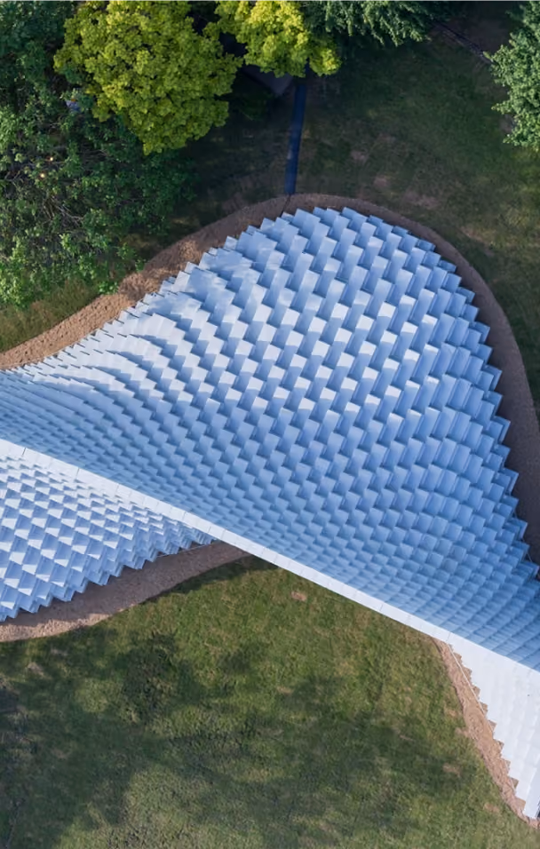 Aerial view of a white, geometric, wave-like architectural structure surrounded by grass and trees.