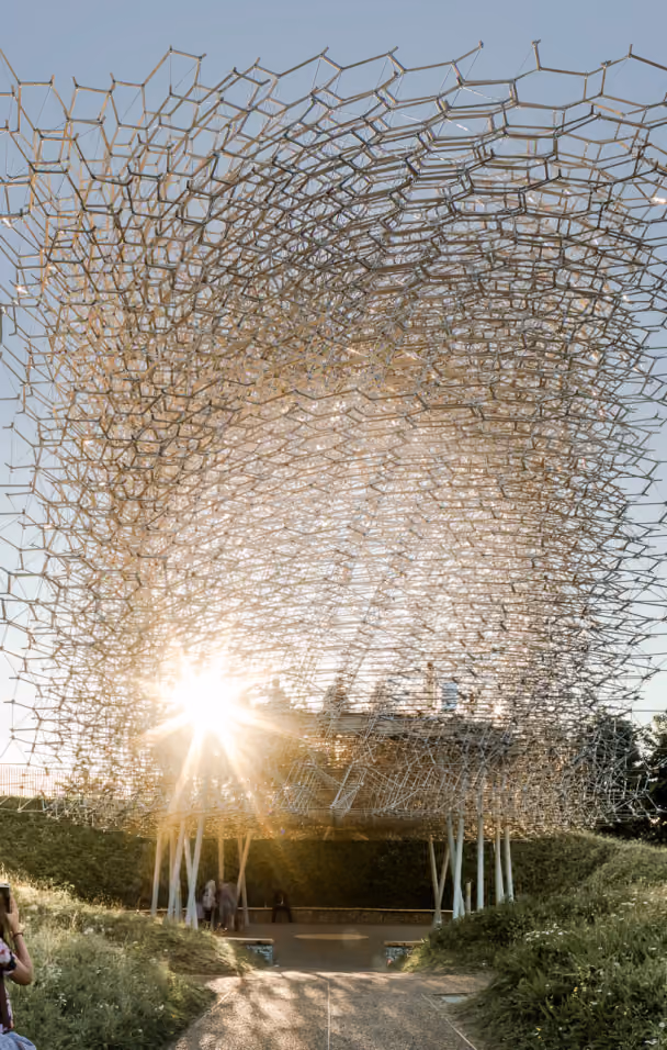 Large transparent honeycomb-like sculpture with sun shining through, supported by white pillars outdoors.