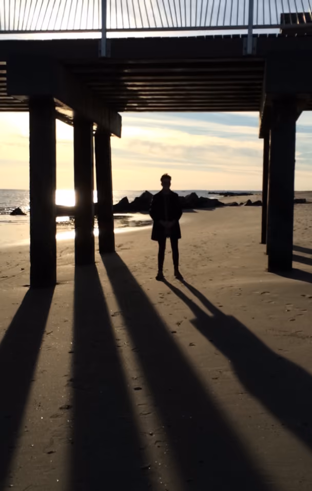 Silhouette of a person standing under a pier casting long shadows on the beach at sunset.