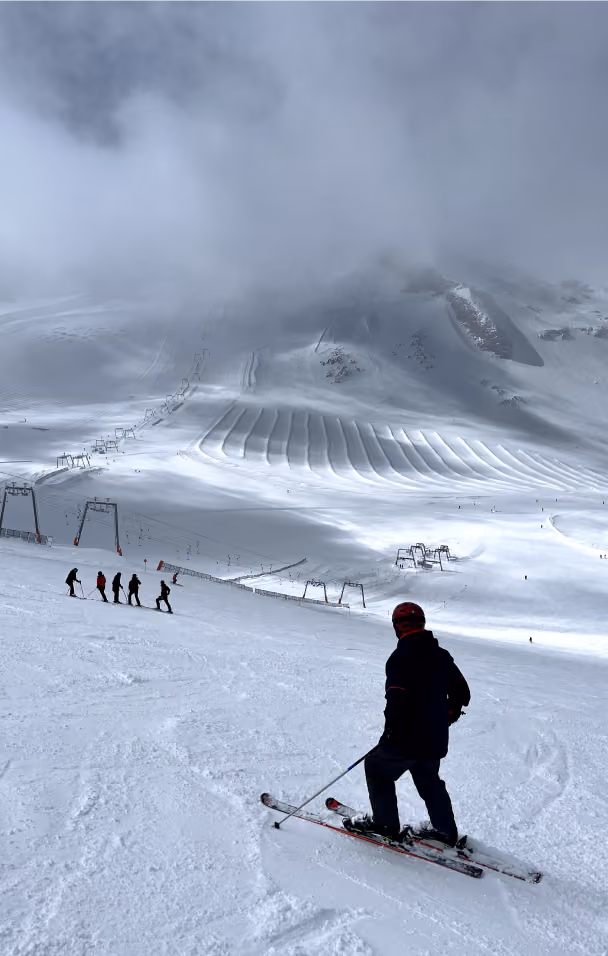 Skier in foreground on snowy slope with group of skiers and groomed trails on mountain under cloudy sky.